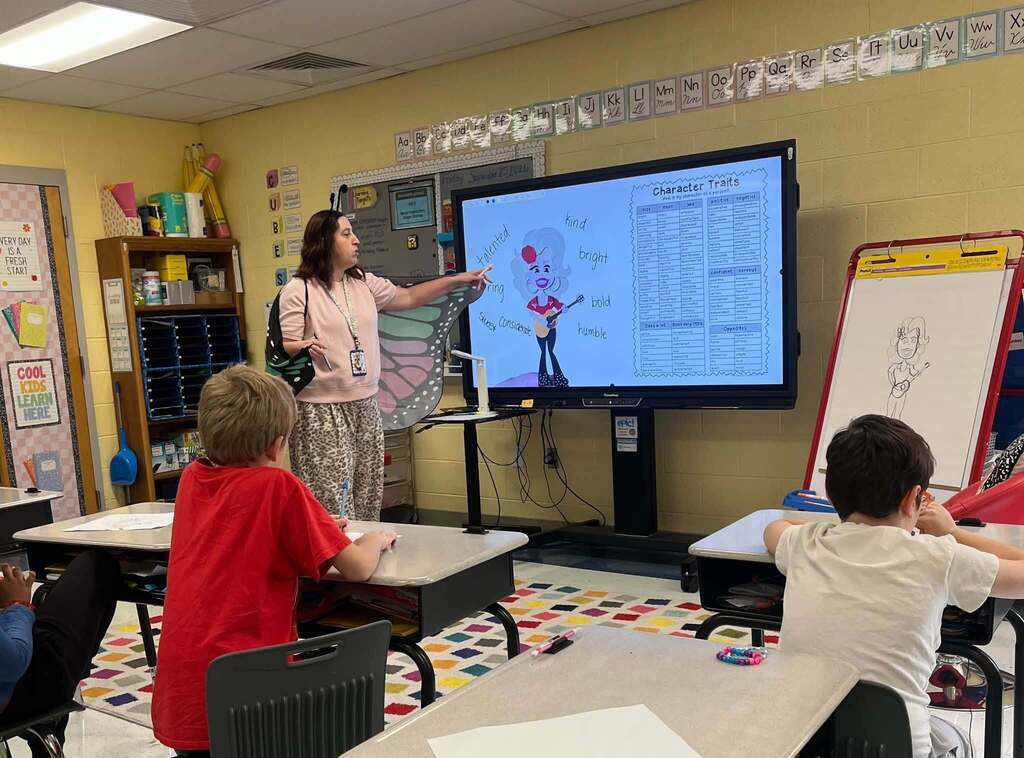 A teacher leads a classroom lesson about Dolly Parton’s character traits while elementary students work at their desks. A Dolly Parton illustration and words including “kind,” “bright,” “bold,” and “humble” are displayed on the classroom screen.