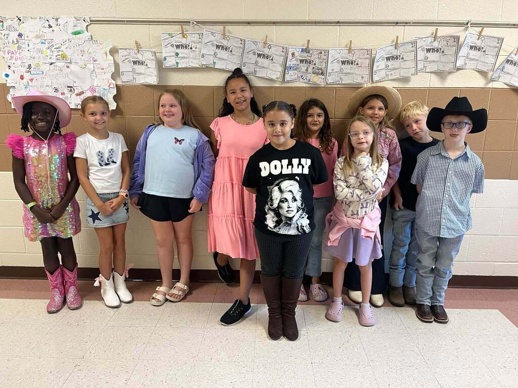 A group of elementary students pose together during Dolly Day in pink, denim, cowboy hats, boots, and Dolly Parton-themed clothing. Student work is displayed on the classroom wall behind them.
