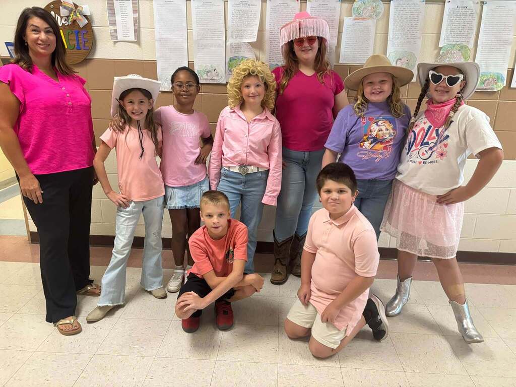Two staff members and a group of elementary students pose together in colorful Dolly Day outfits, including pink clothing, cowboy hats, heart-shaped sunglasses, boots, and a curly blonde wig.