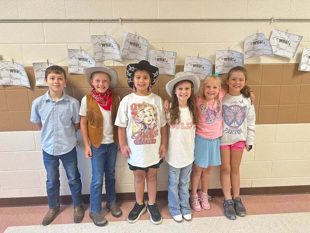 Six elementary students pose together for Dolly Day, with several wearing cowboy hats, boots, pink clothing, and Dolly Parton-themed shirts.