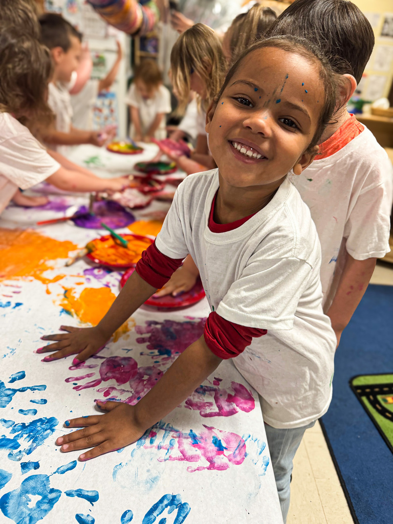 A smiling Pre-K student shows off paint-covered hands during a colorful classroom art activity, with classmates creating handprints in the background.