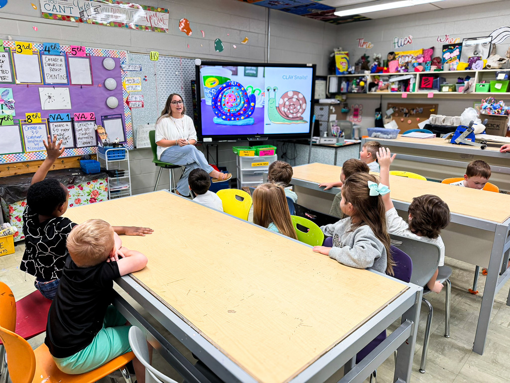 Pre-K students participate in an art lesson, listening to their teacher as she introduces a colorful clay snail project. Several students raise their hands to participate.