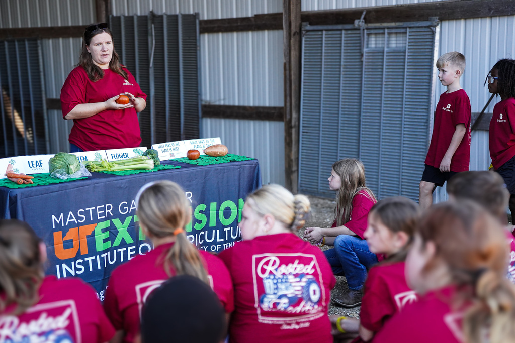 Woman standing behind a table with produce. A group of children and adults in red shirts watch.