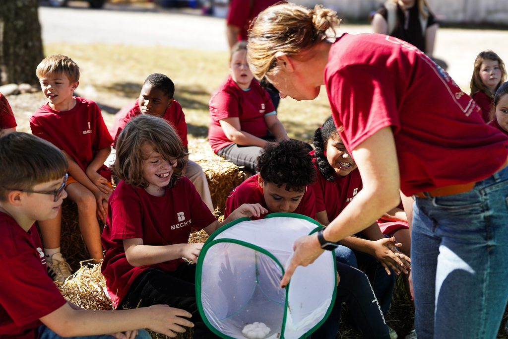 A woman in red holds a net while several children in red shirts look on, seated on hay.