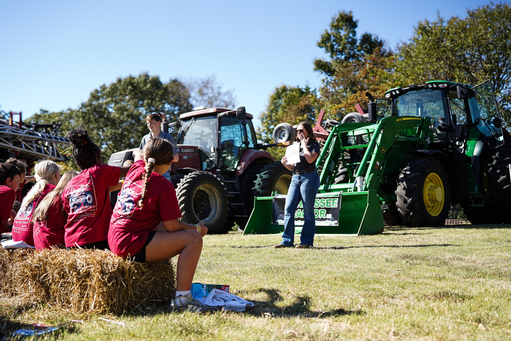 A group sits on hay bales, a person stands in front of a tractor, and a woman speaks.