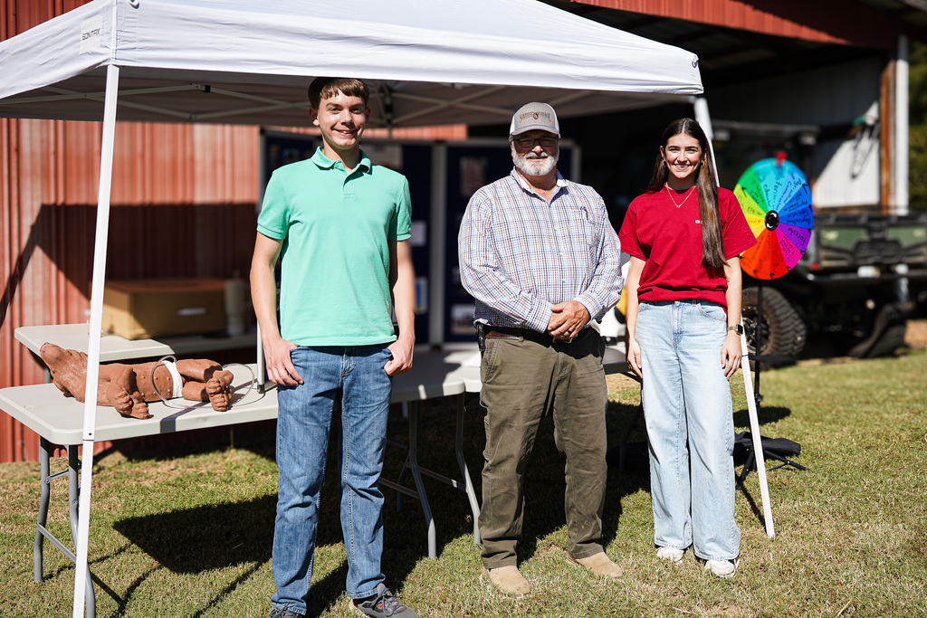 Three people stand under a tent near a red building. A man in green, a man in plaid, and a woman in red.