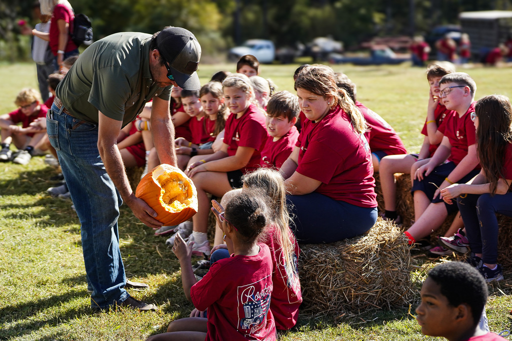 Man showing a pumpkin to seated children outdoors. Children wear red shirts and sit on hay bales.