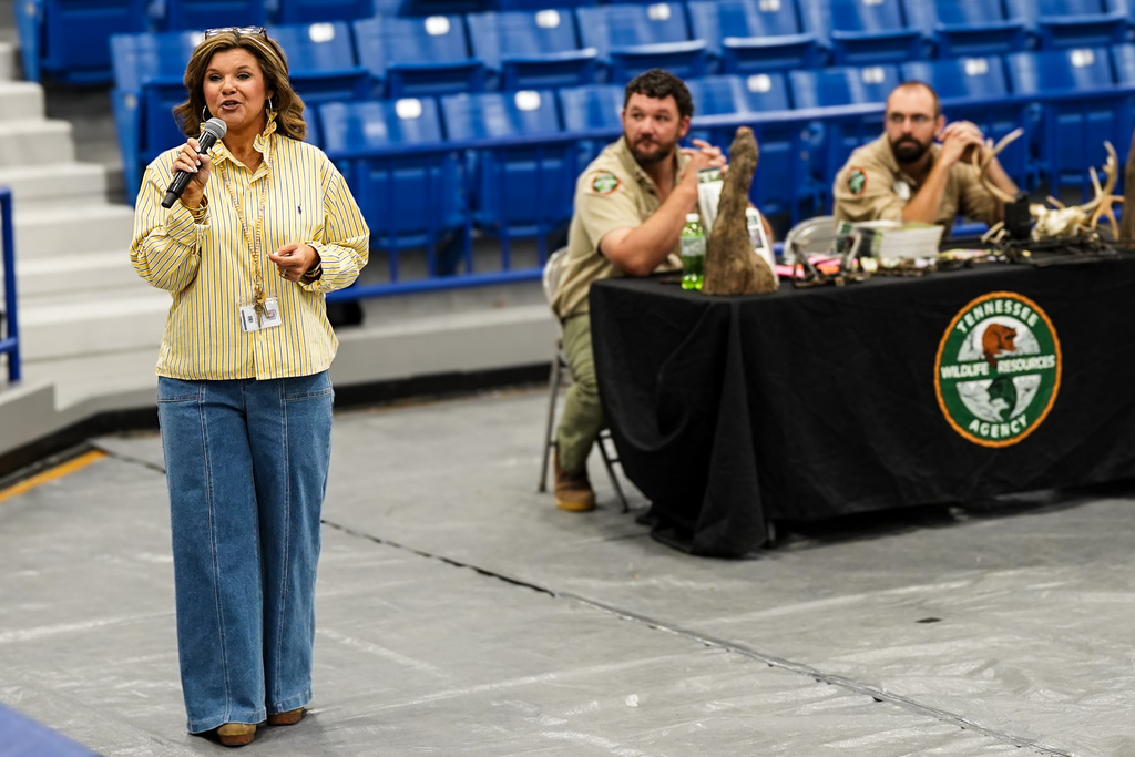 A woman speaks into a microphone, wearing a yellow shirt and blue pants. Two men sit behind a table.