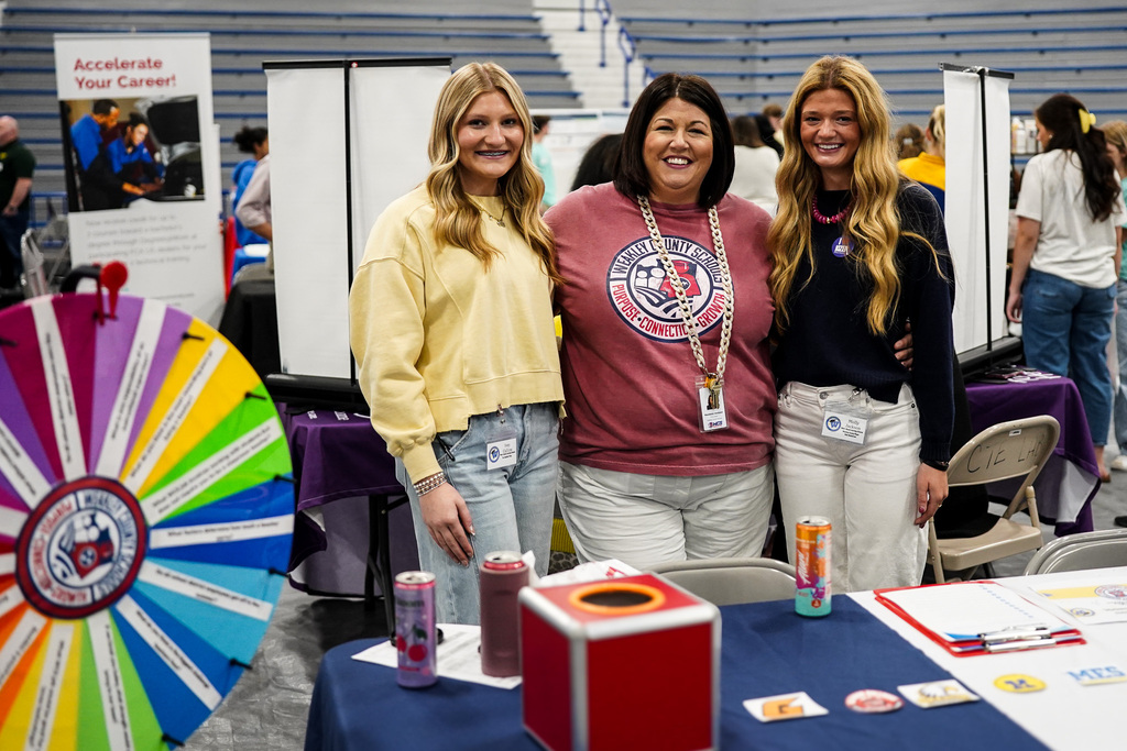 Three women pose near a colorful wheel and table displaying various items, including bottles, a box, and cards.