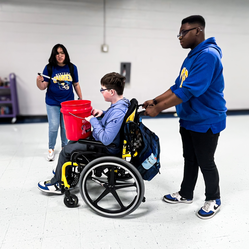 Three Westview High School students participate in a hands-on activity in a school room. One student sits in a wheelchair holding a red bucket, another stands behind the wheelchair, and a third stands nearby holding an item. The students appear to be working together on a task.