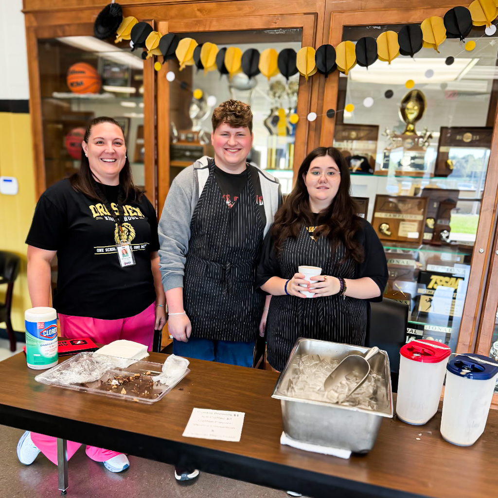 Two Dresden High School Transition students wearing black aprons stand with a staff member behind a table set up for a coffee cart and bake sale. The table holds baked goods, cups, an ice container, and drink supplies, with a decorated school trophy case in the background.