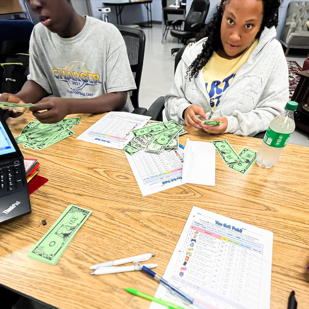 Two Martin Middle School students practice money management skills at a table covered with play money, worksheets, and pencils. Both students are counting and organizing bills as part of a hands-on financial literacy activity.
