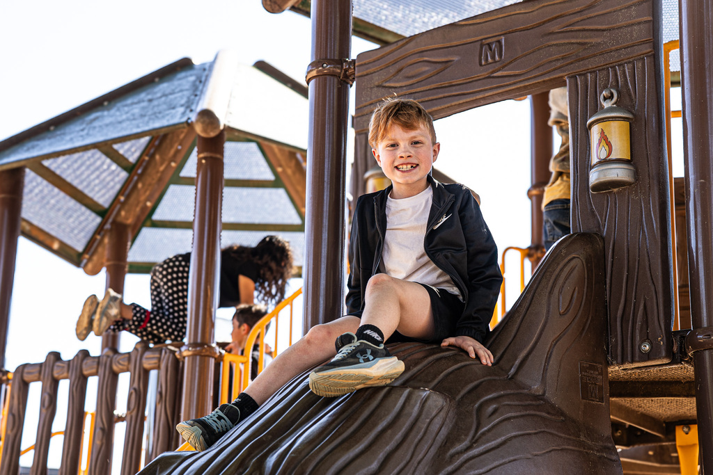 A smiling student wearing a black jacket and shorts sits at the top of a brown playground slide, with other children playing in the background.