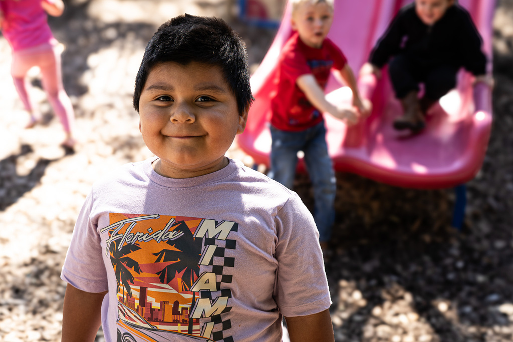 A smiling student wearing a light purple graphic T-shirt stands in front of a playground slide, with other children playing in the background.