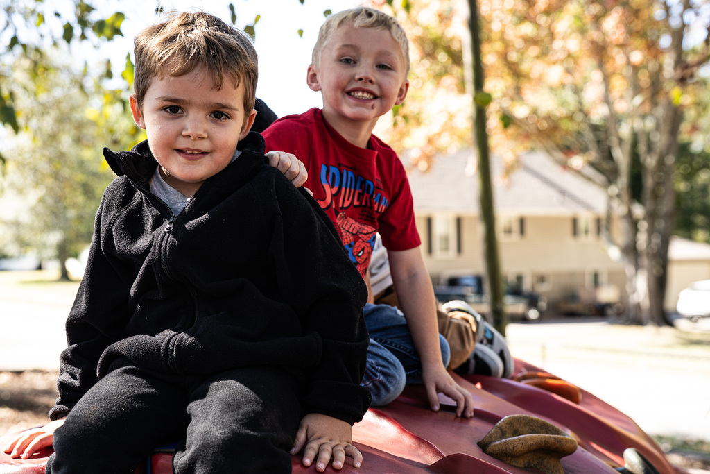 Two smiling students sit together on red playground equipment. One wears a black jacket, while the other wears a red Spider-Man shirt.