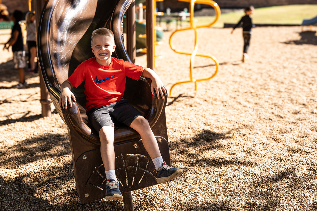 A smiling student wearing a bright red shirt and dark shorts sits at the bottom of a brown playground slide on a sunny day.