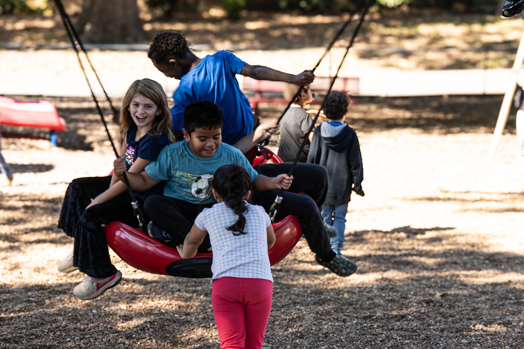 Several students enjoy playground swings together. Three children share a large red circular swing while another student stands nearby, surrounded by other children playing.
