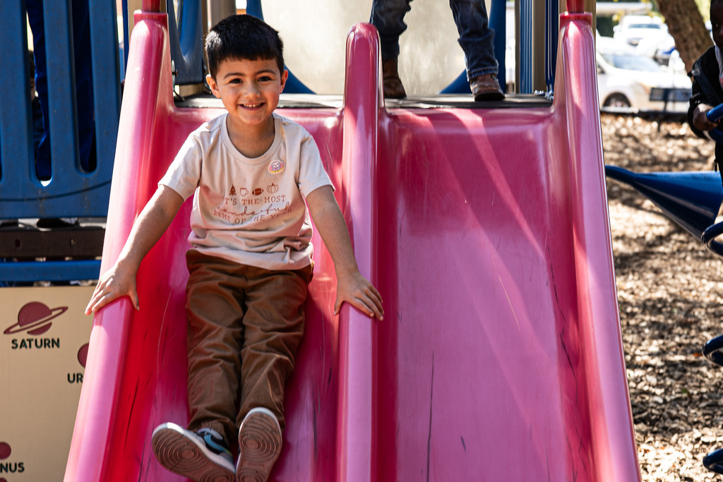 A young student wearing a white T-shirt and brown pants smiles while sliding down a bright pink playground slide.