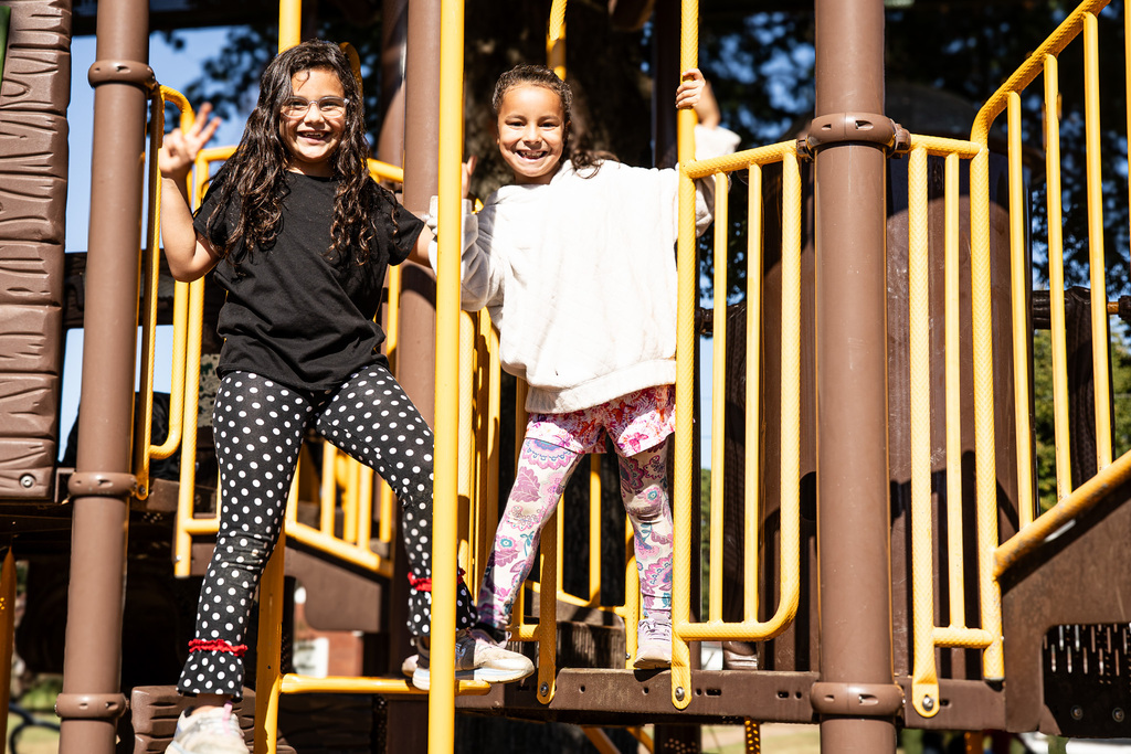 Two smiling students pose together on a playground structure, holding onto bright yellow railings. One wears a black shirt and polka-dot pants, while the other wears a white sweatshirt and colorful patterned leggings.