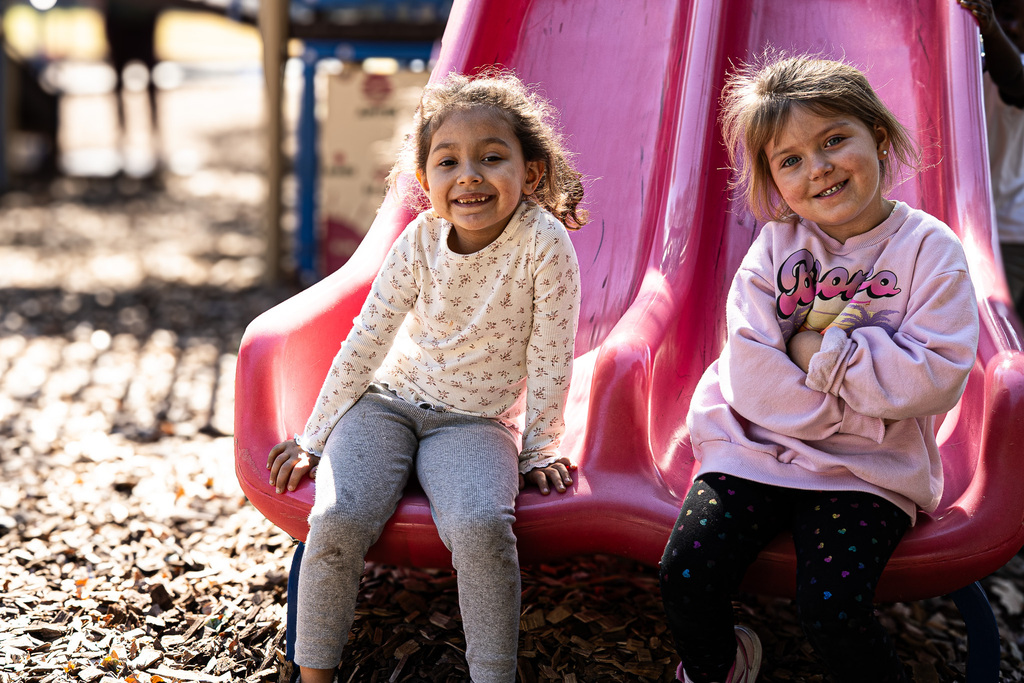 Two smiling students sit side by side at the bottom of a pink playground slide. One wears a cream-colored shirt and gray pants, while the other wears a light pink sweatshirt.