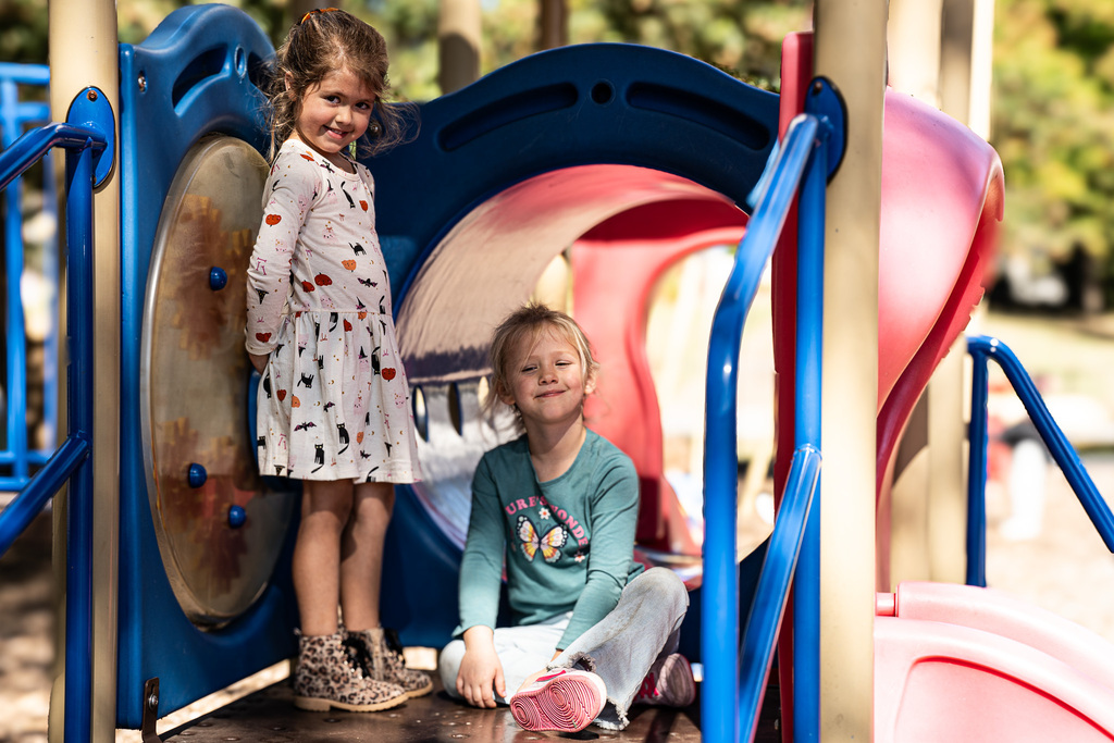wo young students smile for the camera on a colorful playground structure. One stands beside a tunnel opening wearing a white patterned dress, while the other sits nearby wearing a teal shirt.