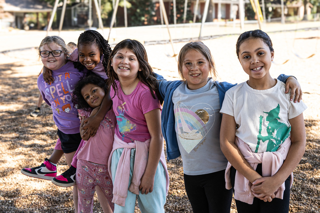 Six smiling students pose together on the playground, standing close with their arms around one another. Swings and playground equipment are visible in the background.