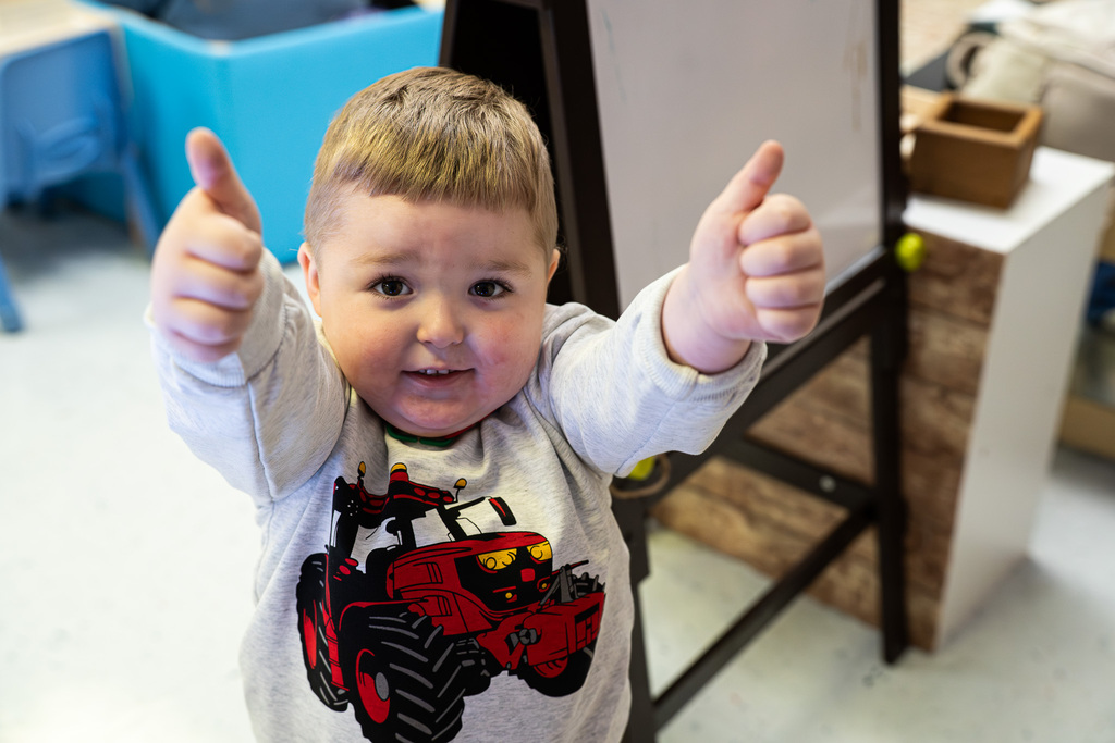 A smiling Pre-K student wearing a tractor sweatshirt gives two thumbs up toward the camera.