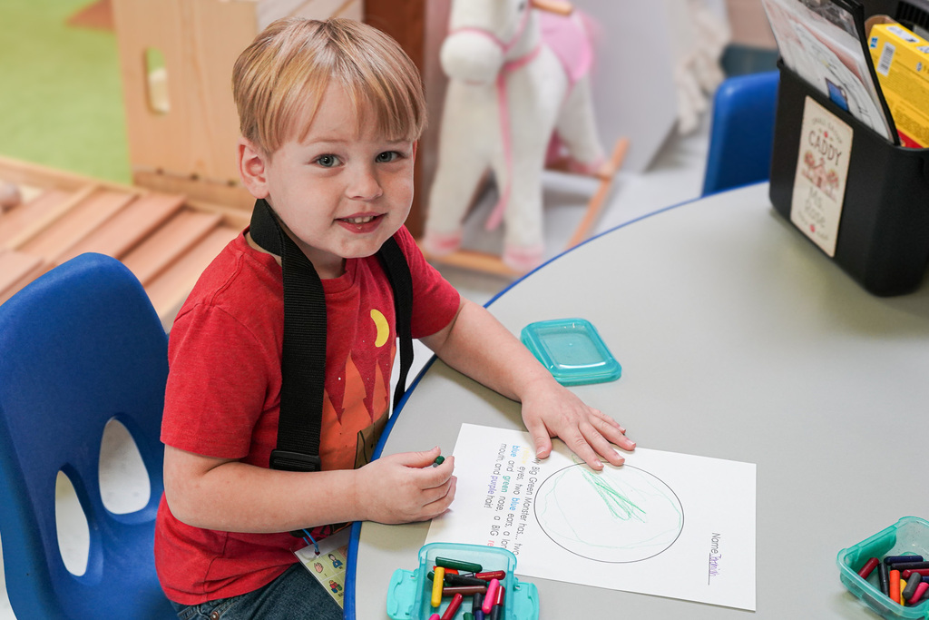 A young student in a red shirt smiles at the camera while coloring a monster-themed worksheet at a classroom table.