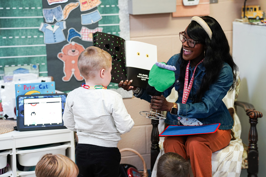 A Pre-K teacher smiles as she uses a green monster puppet and an illustrated book to interact with a student during storytime.
