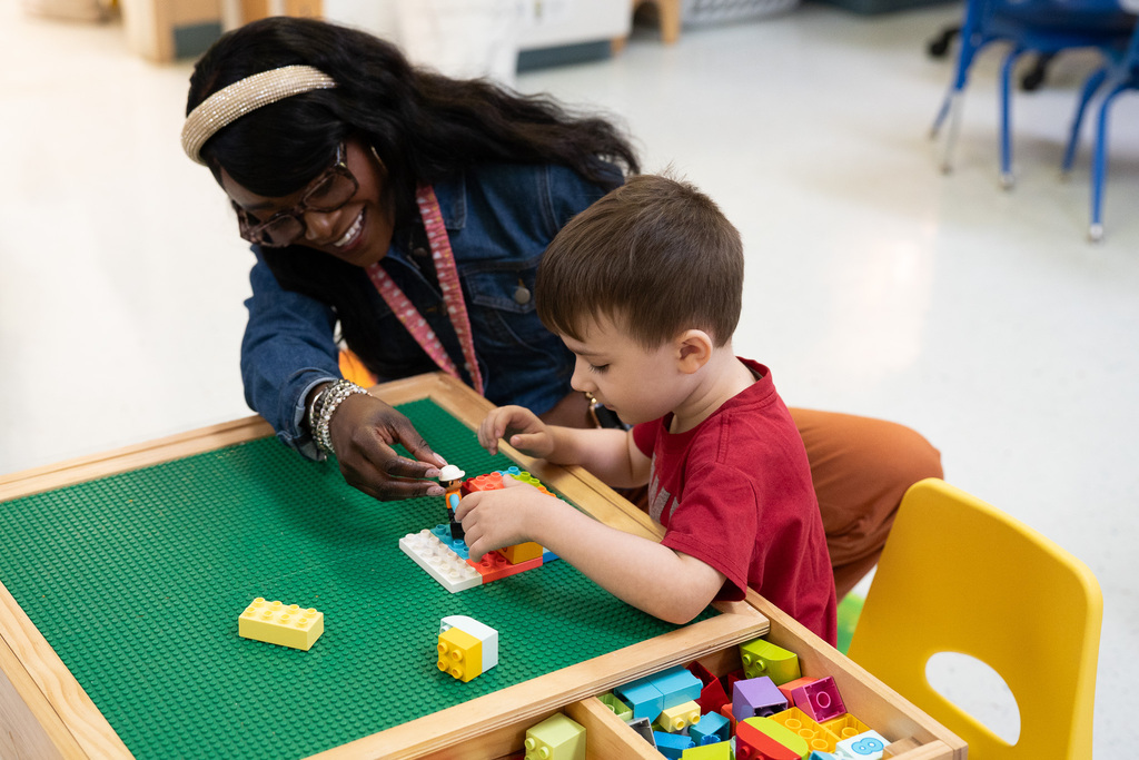 A Pre-K teacher sits beside a young student at a building block table, helping him assemble colorful blocks during playtime.