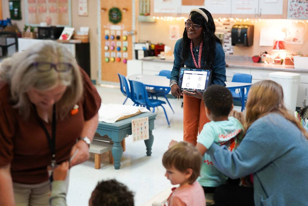 A Pre-K teacher smiles while holding a tablet displaying four letter choices as students and classroom staff participate in a group activity.