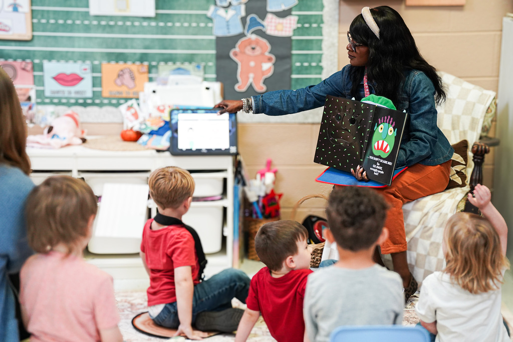 A Pre-K teacher sits in a classroom reading a colorful monster-themed book to a group of young students gathered around her.