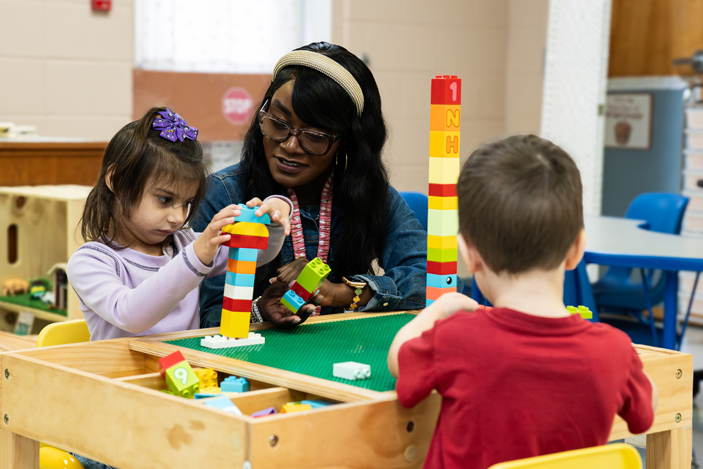 A Pre-K teacher helps two young students build colorful towers with large interlocking blocks during a hands-on classroom activity.