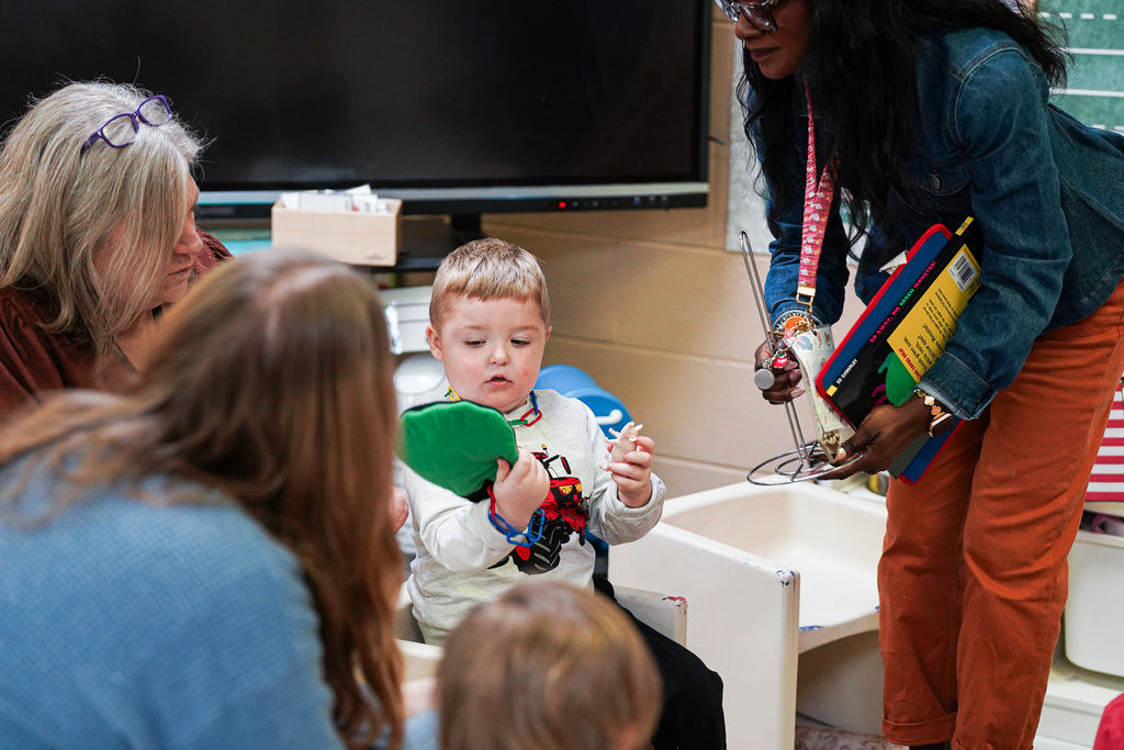 A young student examines a green puppet while two educators sit nearby, supporting him during a classroom activity.