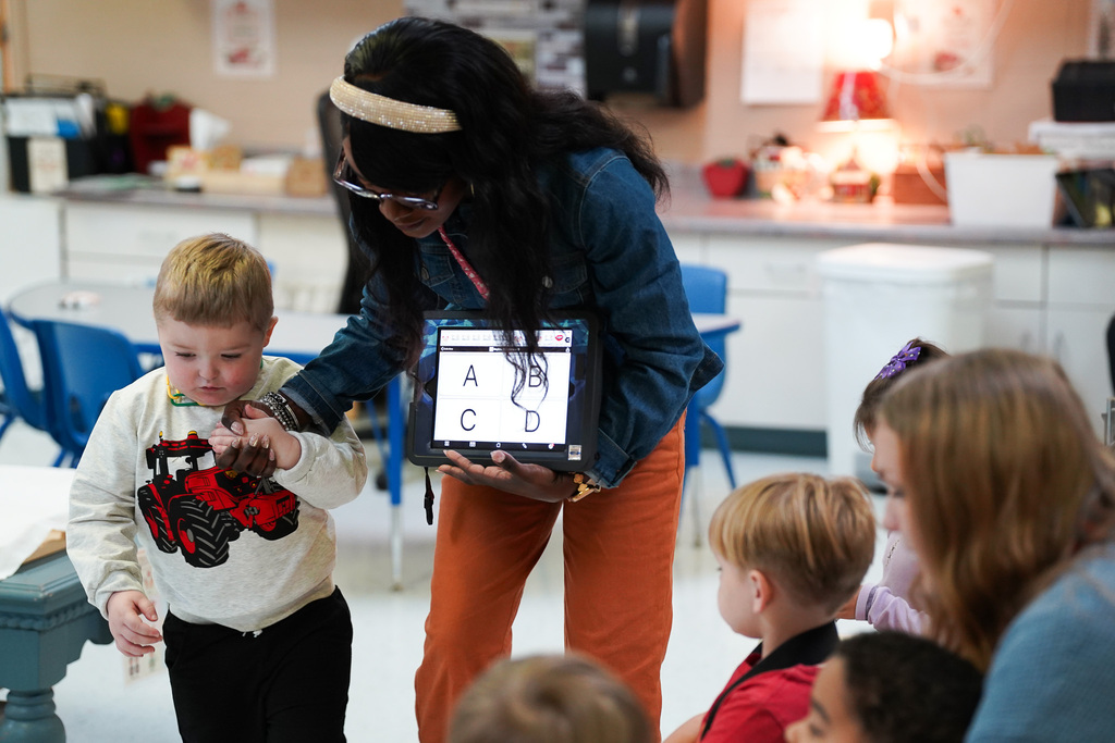 A Pre-K teacher gently supports a student's hand while presenting letter choices on a tablet during an interactive learning activity.