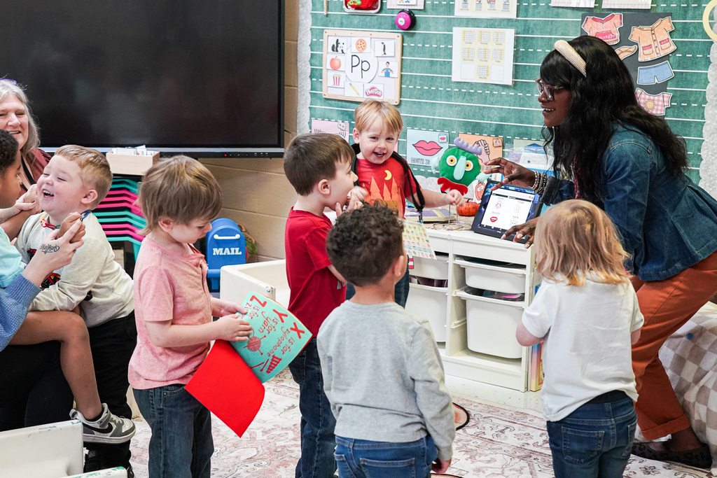 A Pre-K teacher engages a group of young students using a green monster puppet and a tablet displaying communication symbols.