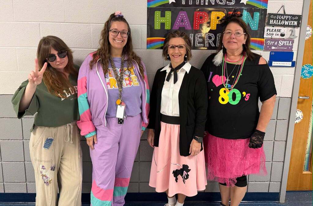 Four female staff members pose in a school hallway wearing outfits inspired by different decades, including a 1970s peace-sign look, a colorful 1990s tracksuit, a 1950s poodle skirt, and bright 1980s accessories for Red Ribbon Week.