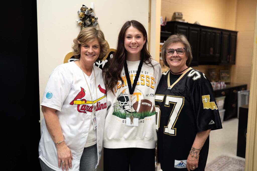 Three female staff members smile together wearing sports apparel representing the St. Louis Cardinals, Tennessee Volunteers, and New Orleans Saints for Red Ribbon Week.