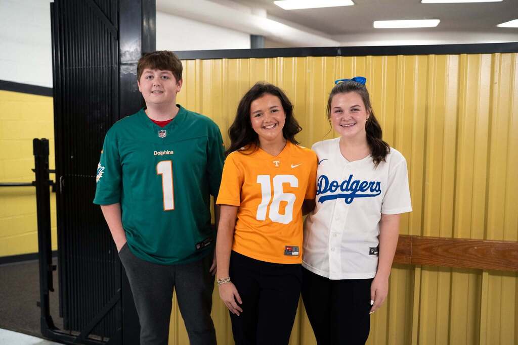 Three high school students stand together in front of a black and gold wall wearing Miami Dolphins, Tennessee Volunteers, and Los Angeles Dodgers jerseys for Red Ribbon Week.