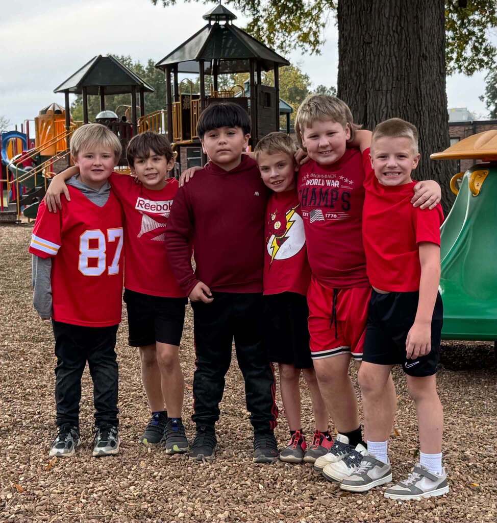 Six elementary students stand together with their arms around one another on a school playground, wearing red shirts, sweatshirts, and sports jerseys for Red Ribbon Week.