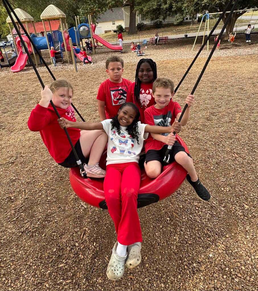 Five young students gather on a large red playground swing, with several wearing red clothing in celebration of Red Ribbon Week. Other children play on the playground in the background.