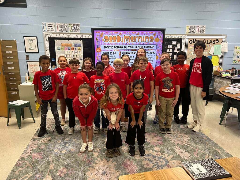 A group of elementary students and their teacher pose inside a classroom wearing red shirts and sweatshirts to participate in Red Ribbon Week.