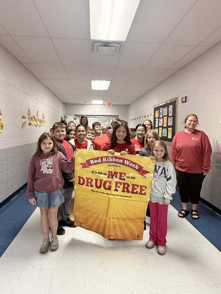 A group of elementary students and staff members gather in a school hallway, with several students holding a large yellow Red Ribbon Week banner reading, "It's Up to ME to Be Drug Free."