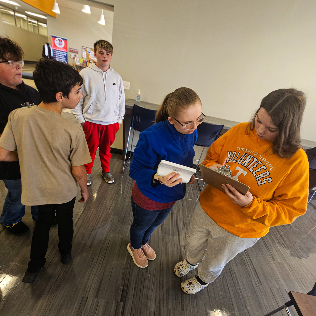 A student in a blue jacket examines readings on a handheld air-quality monitor while another student records information on a clipboard. Three classmates observe nearby as part of the Transition Program's air-quality testing project.