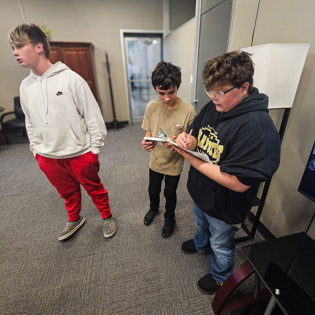 Three students in the Weakley County Transition Program stand inside an office area. One student writes on a clipboard while another holds paperwork, documenting information during the district's air-quality testing project funded by a TVA grant.