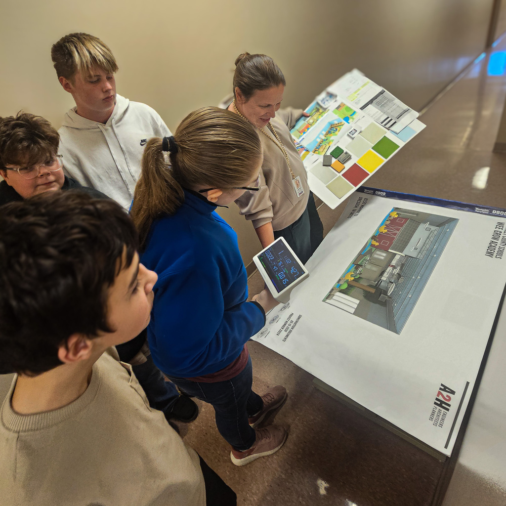 Students in the Weakley County Transition Program gather around architectural plans and color samples while a student in a blue jacket holds a digital air-quality monitor during a visit to a district building.