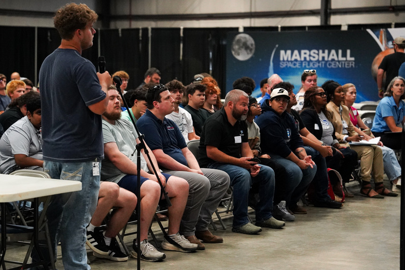 A student stands at a microphone to ask a question during a presentation at NASA’s Marshall Space Flight Center as other students listen.