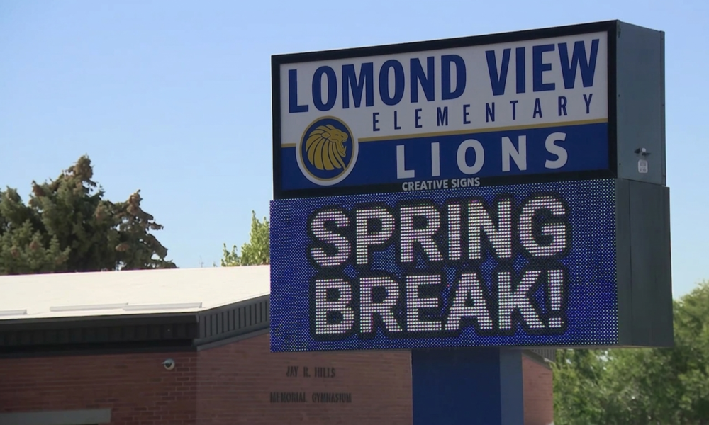 An image of a large sign for "Lomond View Elementary Lions," featuring a graphic of a lion's head, below which an electronic LED reader board displays the message "SPRING BREAK!". The sign stands under a clear blue sky, with a red brick building visible at the base and various green trees and foliage surrounding the area.