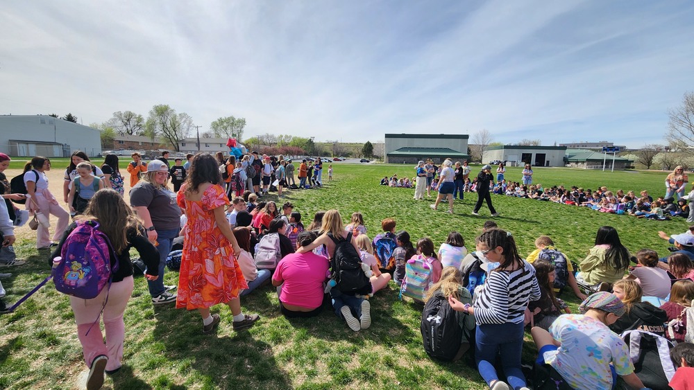 A group of teachers and students sitting on the grass outside with buildings in the background and bright blue sky