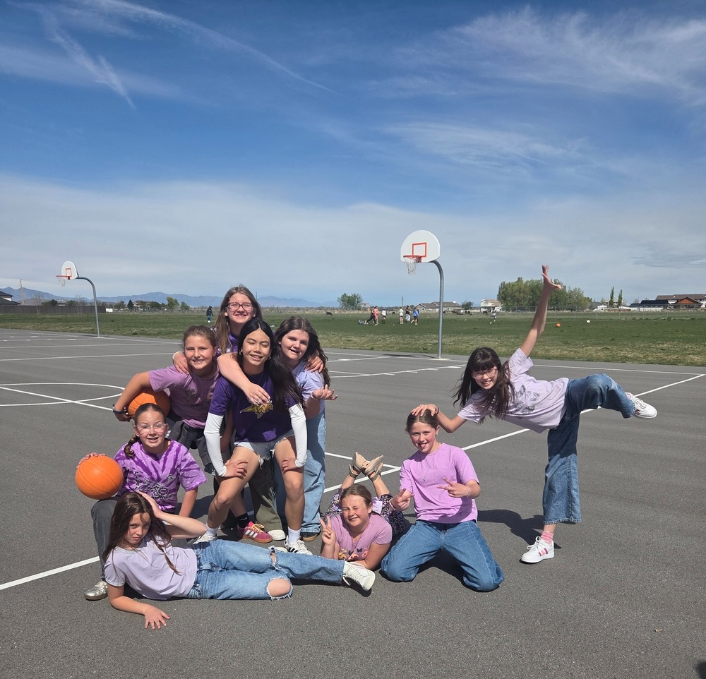 A group of nine smiling girls poses playfully on an outdoor asphalt basketball court under a clear blue sky with light, wispy clouds. The group is arranged in a dynamic, multi-level composition: Left Side: One girl reclines on the ground in front, while another sits behind her holding a basketball. Two more girls stand behind them, one wearing a purple tie-dye shirt and the other a solid purple shirt. Center: A girl in a purple t-shirt with a yellow graphic stands prominently, surrounded by two friends with their arms around her. Another girl lies on her stomach on the ground in front of them, kicking her feet up. Right Side: One girl kneels while making peace signs, and another stands on one leg in a "star" pose with her arms outstretched. The background features two basketball hoops, a green grassy field, and distant mountains under a bright horizon. There is no visible text or writing on the clothing or surroundings other than the faint graphic on the center girl's shirt.